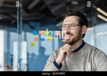 Selbstbewusster bärtiger Mann mit einer Brille, die lächelt, wegblickt und Hand am Kinn hält, repräsentiert Geschäftserfolg, innovative Ideen und durchdachte Planung in einer modernen Büroumgebung Stockfoto