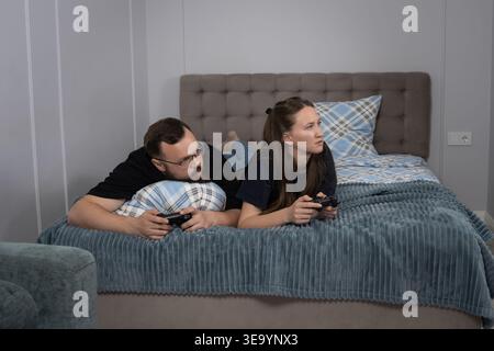 Junge Paare liegen auf einem Bett, konzentriert und kompetitiv, während sie Videospiele mit Controllern spielen, entspanntes Home Entertainment, Bonding und Shar genießen Stockfoto
