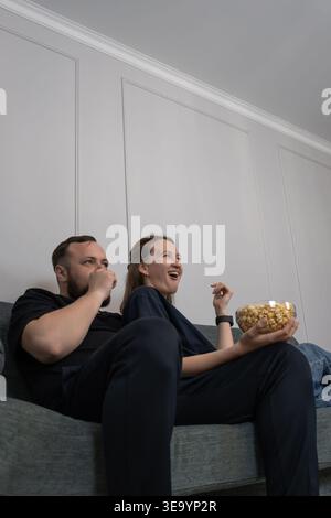 Paare entspannen sich abends auf einem Sofa zu Hause, teilen Popcorn und sehen sich gemeinsam einen Film an, gemütliche zwanglose Momente der Verbindung und ruhige Wochenendeinfahrt Stockfoto