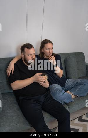 Junge Paare auf Sofa, die fernsehen und Popcorn essen; Frau reagiert mit Ekel, während der Mann weiter schnappt, freimütige häusliche Szene mit kontrastierendem Emotion Stockfoto