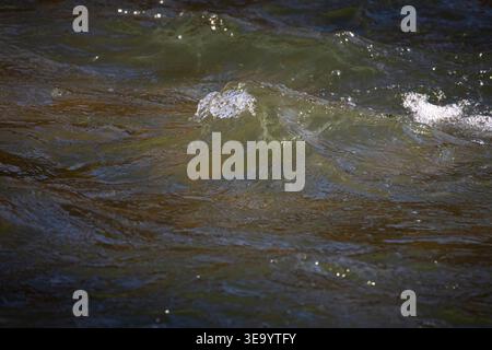 Aus nächster Nähe sehen Sie das Flusswasser in Bewegung mit kleinen Wellen und hellen Blasen, die über dunkelgrünen Strömungen aufragen, subtile Reflexionen und Kräuselungen Stockfoto