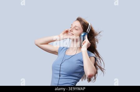 Halblanges Porträt einer jungen Frau, die Kopfhörer trägt, die Augen geschlossen und lächelnd, die Haare vom Wind geweht, Musik in einer unbeschwerten Lifestyle-Mome genießt Stockfoto