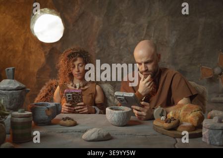 Prähistorische Paare, die zu Hause frühstücken, benutzen ihre Smartphones und ignorieren einander Stockfoto