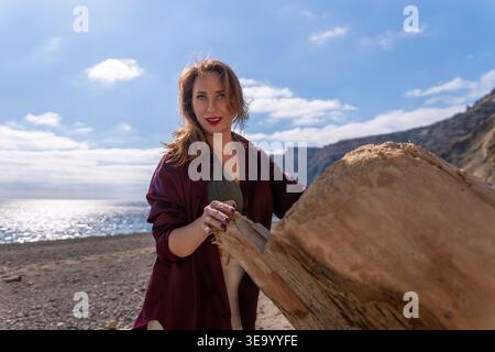 Woman Beach Treibholz steht hinter einem großen Stück Holz an einem sonnigen Kieselstrand unter einem klaren blauen Himmel Stockfoto