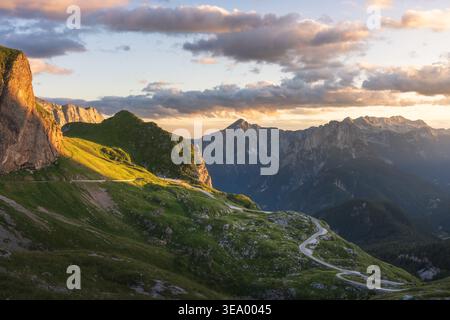 Blick aus der Vogelperspektive auf goldenes Sonnenlicht, das die zerklüfteten Gipfel und Serpentinenstraßen küsst, die sich durch grüne Hänge schlängeln, Log Pod Mangartom, Bovec, Slowenien. Stockfoto