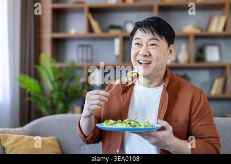 Asiatischer Mann genießt einen gesunden grünen Salat von einem blauen Teller, während er auf einer Couch in einer gemütlichen häuslichen Umgebung sitzt und Wohlbefinden und eine bewusste Ernährung fördert Stockfoto