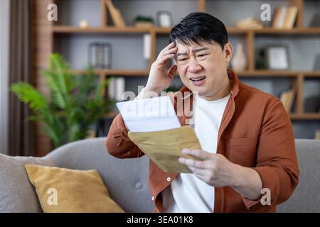 Gestresster asiatischer Mann, der auf einem Sofa in einer modernen Wohnung sitzt, einen Umschlag mit einem Brief in der Hand hält und den Kopf mit der Hand bedeckt und auf schlechte Nachrichten reagiert Stockfoto