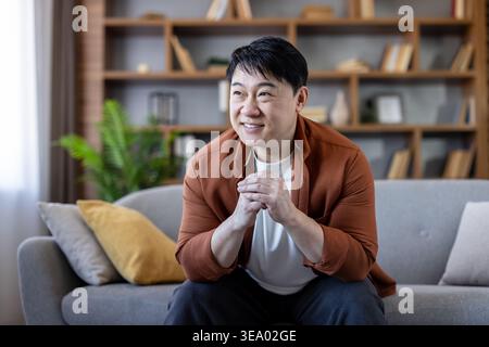 asiatischer Mann mittleren Alters, der auf einem bequemen Sofa in einem modernen Wohnzimmer lächelt und entspannt, die Hände gefesselt und friedlich zwischen gemütlichen Kissen und Bücherregalen wegblickt Stockfoto