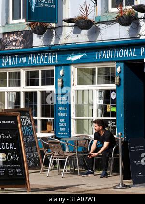 Ein junger Trinker sitzt vor dem Sprott and Mackerel Pub in der malerischen Küstenstadt Brixham, Devon, Frühjahr 2026 Stockfoto