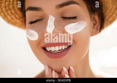 Frau lächelt mit Sonnencreme auf den Wangen und Nase berührend das Kinn. Junge Frau mit Strohhut. Weibliches Modell, das Sonnenlicht genießt. Hautpflege und Sonnenschutz Stockfoto