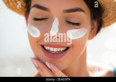 Frau lächelt mit Sonnencreme im Gesicht und berührt die Haut mit beiden Händen. Junge Frau, die Sonnenlicht genießt. Weibliches Modell mit Hautpflegeroutine. Sonnenschutz Stockfoto
