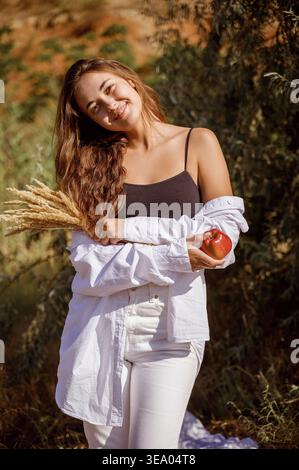 Junge Frau, die auf sonnigem Feld steht und rote Äpfel und getrocknetes Gras hält Stockfoto