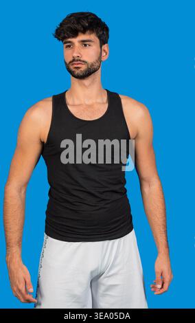 Junger Mann, der in einem Tanktop und Shorts vor blauem Hintergrund steht Stockfoto
