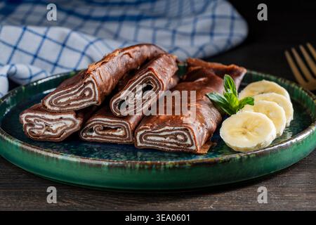 Köstliche Schokoladenpfannkuchen, gerollt mit Sahnefüllung, serviert mit frischen Bananenscheiben und Minze auf grünem Teller, Nahaufnahme. Perfekt für Dessertmenü, Stockfoto