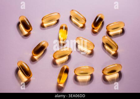 Goldene flüssige Gelkapseln auf violettem Hintergrund. Durchscheinendes Bernsteinfischöl oder Vitaminweichgel auf mattem Flieder mit dramatischem shado Stockfoto