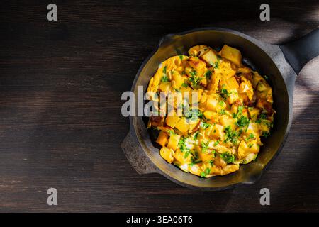 Gebratene Kartoffeln mit Eiern und Petersilie in der Pfanne. Hausgemachte Kartoffelkartoffeln mit Rührei und frischen Kräutern in schwarzer Pfanne auf dunklem Holz. Rustikaler Urlaub Stockfoto