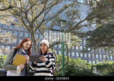 Weibliche Freunde, die in der Nähe von Laternenpfosten auf dem Campus stehen, Tablette und gelbe Mappe halten und eine Mütze tragen. Outdoor, Urban, plaza, Grünflächen, Universität, Studie, Paar Stockfoto