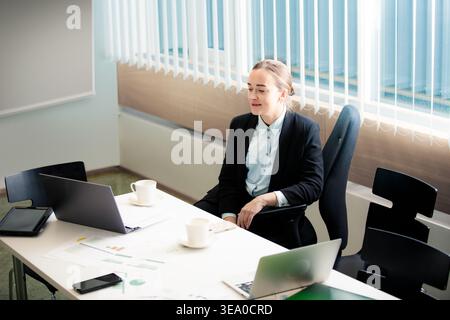 Professionelle Geschäftsfrau, die in einem hellen Büro an einem Laptop arbeitet. Konzentrierte Mitarbeiterin analysiert Berichte mit einer Tasse Kaffee. Stockfoto