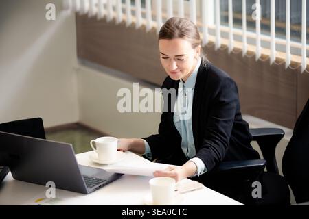 Professionelle Geschäftsfrau, die in einem hellen Büro an einem Laptop arbeitet. Konzentrierte Mitarbeiterin analysiert Berichte mit einer Tasse Kaffee. Stockfoto