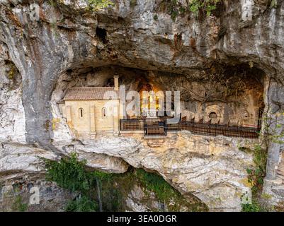 Aus der Vogelperspektive auf den Santuario de Covadonga, der sich am Berghang festhält, ein Leuchtfeuer vor der zerklüfteten, natürlichen Höhle Santa Cueva de Covadonga, Stockfoto