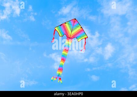 Farbenfroher Regenbogendrachen, der hoch in einem hellblauen Sommerhimmel mit weichen weißen Wolken aufsteigt und Freiheit, Kinderfreude und Outdoor-Fu symbolisiert Stockfoto