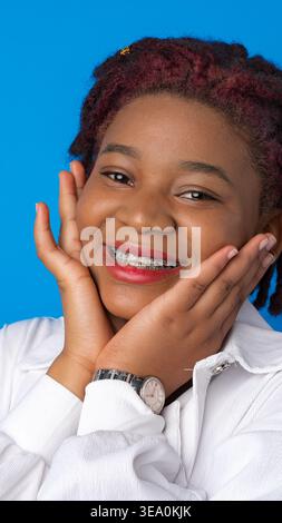 Teenager lächelt, während sie ihr Gesicht mit beiden Händen vor blauem Hintergrund hält Stockfoto