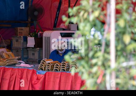 Ein Lebensmittelverkäufer mit Gesichtsmaske und Haarnetz nimmt an einem Marktstand im Freien in Penang, Malaysia, gebackenes Gebäck an. Eingerahmt durch tropisches Laub, fängt die Szene einen authentischen Blick auf die lokale Zubereitung von Speisen ein. Stockfoto