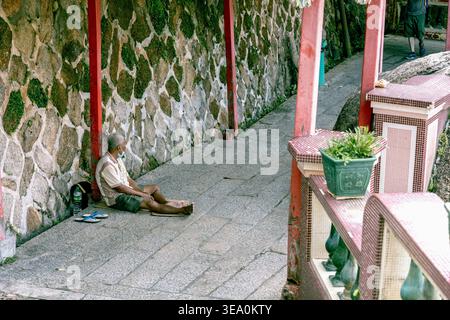 Ein älterer Mann ruht auf dem Boden vor einer Granitsteinmauer in einem Flur des Kek Lok Si Tempels in Penang, Malaysia. Die offene Szene fängt einen ruhigen Moment der Einsamkeit zwischen den roten Säulen und Mosaikgeländern eines der größten buddhistischen Tempel Südostasiens ein. Stockfoto