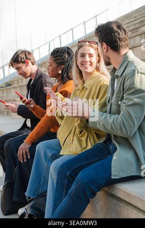 Eine fröhliche Gruppe von Freunden, die gerne interagieren und ihr Smartphone nutzen, während sie auf den Stufen sitzen, Momente teilen und lachen, was eine Freude schafft Stockfoto