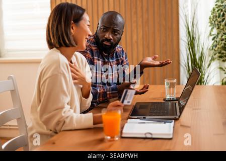 Eine Frau hält eine Kreditkarte, während ihr Partner mit offenen Händen Gesten macht, die beide ernsthaft über Online-Zahlungen sprechen. Stockfoto