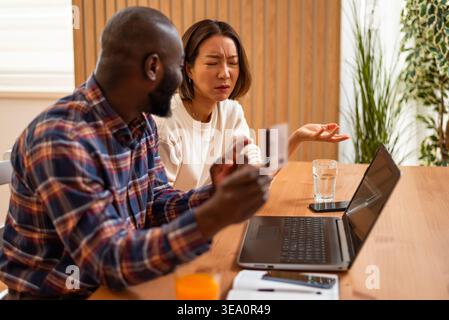 Ein Mann hält eine Kreditkarte, während sein Partner mit offenen Händen Gesten zeigt, beide in einem ernsthaften Gespräch über Online-Zahlungen. Stockfoto