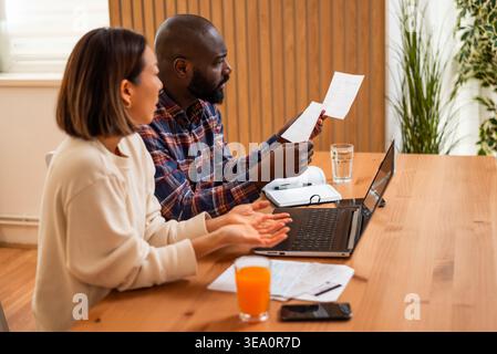 Ein Mann untersucht die Papiere genau, während sein Partner besorgt reagiert. Beide schauen sich einen Laptop an, während er die Haushaltsfinanzen verwaltet. Stockfoto