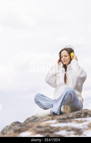 Junge Frau, die Musik genießt, während sie in den Bergen mit kreativen Blumen-Kopfhörern unterwegs ist. Minimaler Außenbereich mit Kopierraum, der Freiheit zum Ausdruck bringt Stockfoto