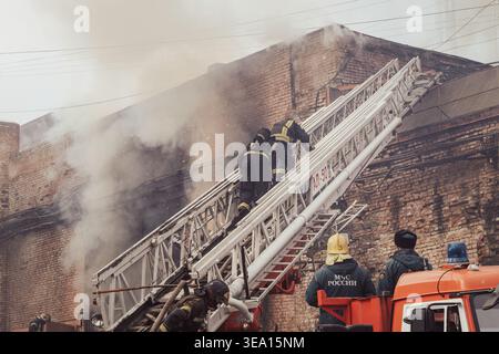 Krasnojarsk, Russland, 14 04 2026: Feuerwehrleute von EMERCOM of Russia (MCHS) nutzen einen Luftleiter-Lkw, um einen Brand in einem Industriegebäude zu bekämpfen. Dick Stockfoto