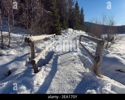 Entlang des verschneiten Weges überquert eine Holzbrücke einen kleinen gefrorenen Bach Stockfoto