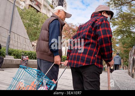 Taipei, Taiwan - 18. Januar 2025: Ein älterer Mann sieht mit einem Stock neben seiner Frau einen gemeinsamen Lebensmittelwagen, der auf einem offenen, winterlichen Bürgersteig in Taipeh zu sehen ist. Stockfoto