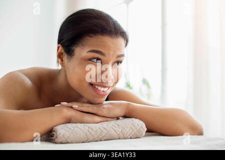 Schwarze Frau lächelt und ruht Kopf auf gefaltetem Handtuch nach Spa-Behandlung Stockfoto