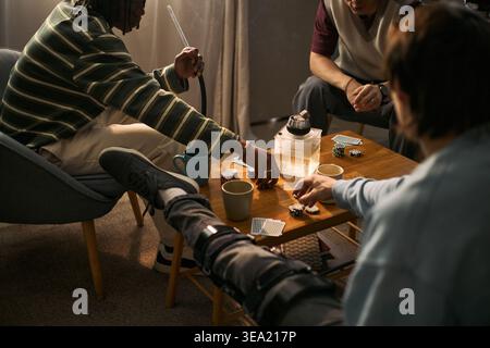 Multiethnische junge Erwachsene spielen Karten und Pokerchips um den Couchtisch und treffen sich während der Heimfeier. Nützlich für Freundschaftskampagnen, Nachtleben, Freizeit und Suchtrisiko Stockfoto
