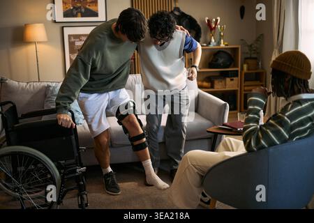 Junger erwachsener Mann mit Behinderung, der neben dem Rollstuhl assistiertes Gehen praktiziert, während er sich auf die Schulter seiner Freunde lehnt, unterstützt Bewegung, nützlich für Rehabilitation, Erholung, Gesundheitsmarketing Stockfoto