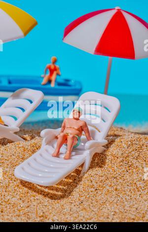 Nahaufnahme eines Miniaturmannes, der auf einer weißen Liege unter einem rot-weißen Sonnenschirm auf Sand liegt, mit einer weiteren Liege und einem Boot mit Figuren Stockfoto