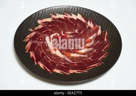 Dünne Stücke köstlicher iberischer Jamon auf einem dunklen Teller mit traditioneller spanischer Küche auf weißem Hintergrund Stockfoto