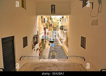Eine Hochwinkelperspektive mit Blick über eine Treppe und durch einen Kalksteinbogen in eine typische enge Straße von Valletta, die Tradition zeigt Stockfoto