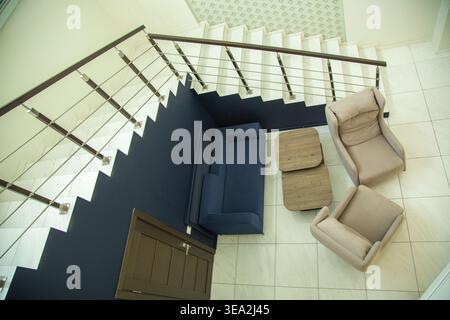 Hochwinkelaufnahme einer modernen Inneneinrichtung mit blauem Sofa, zwei beigefarbenen Sesseln und einem Holztisch neben einer weißen Treppe mit dunklem W Stockfoto