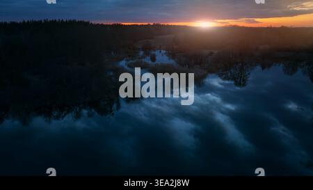 Die Drohnenansicht fängt die ruhige Schönheit eines Sees ein und reflektiert dramatische Dämmerungswolken bei einem lebhaften Sonnenuntergang über einem dichten Wald Stockfoto