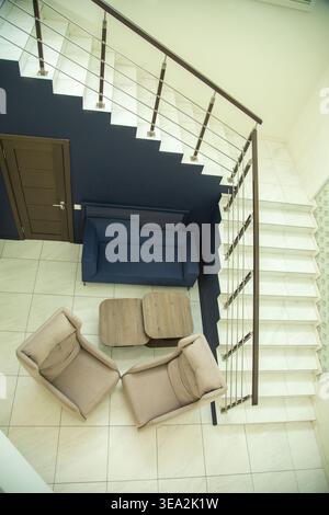 Hochwinkelaufnahme einer modernen Inneneinrichtung mit blauem Sofa, zwei beigefarbenen Sesseln und einem Holztisch neben einer weißen Treppe mit dunklem W Stockfoto