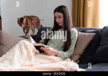 Eine junge Frau, die zu Hause ein Buch liest, in eine kuschelige Decke mit einer bengalischen Katze gewickelt. Warme Indoor-Lifestyle-Szene mit ruhiger Atmosphäre, Entspannung, Hygge m Stockfoto