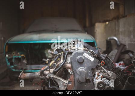 Ein altes, blaues Auto sitzt zerlegt in einer schwach beleuchteten Garage, mit einem Motor im Vordergrund. Stockfoto