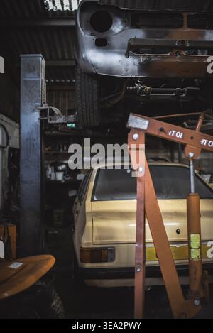 In einer Garage wird an einem alten Auto gearbeitet, dessen Vorderseite angehoben ist, und darunter befindet sich ein gelbes Auto. Stockfoto