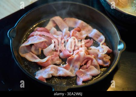 Brutzende Speckstreifen, die in einer gusseisernen Pfanne auf einem Herd braten. Nahaufnahme mit fettigem Speck, der zum Frühstück gekocht wird. Stockfoto