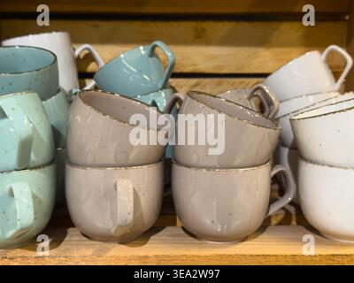 Gestapelte Keramikkkaffeetassen in verschiedenen Pastellfarben auf einem Holzregal. Töpferbecher im rustikalen Stil für Tee oder Kaffee in einem Geschäft. Stockfoto